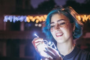 Retrato noturno de close-up de uma jovem sorridente com cabelo azul e óculos de aro redondo, segurando e sendo iluminada por um fio de luzes de LED brancas.
