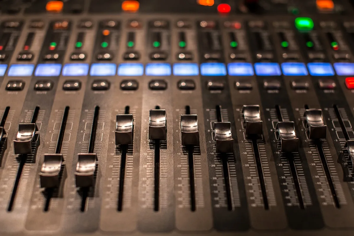 Mesa de som profissional com vários faders prateados ajustando volumes e botões iluminados em azul, verde e vermelho.