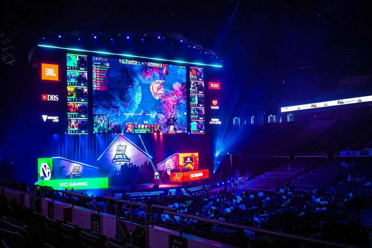 Telão de LED gigante em um evento de E-sports, exibindo a transmissão do jogo e placares, com o público na arquibancada escura, representando a tecnologia em grandes competições de jogos.