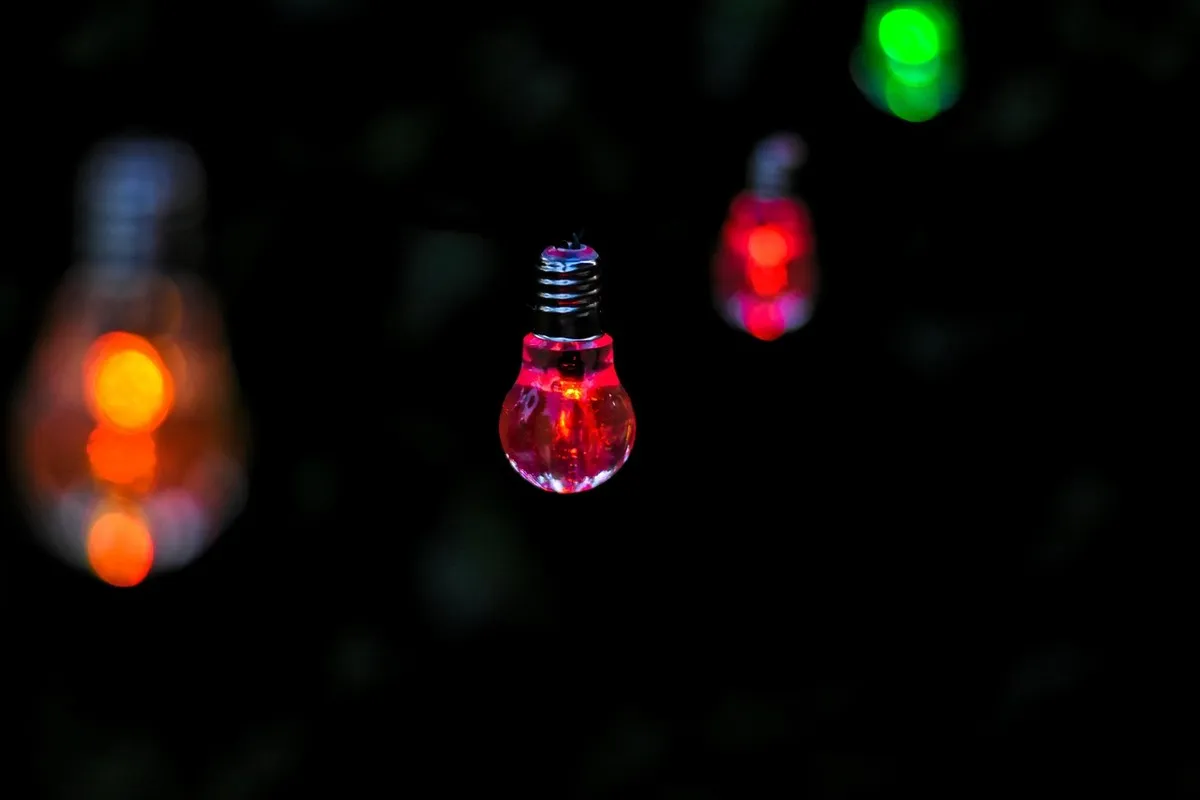 Close-up em um ambiente escuro de uma pequena lâmpada decorativa com rosca (possivelmente LED), brilhando em luz vermelha intensa, com outras luzes coloridas desfocadas (bokeh) ao fundo.