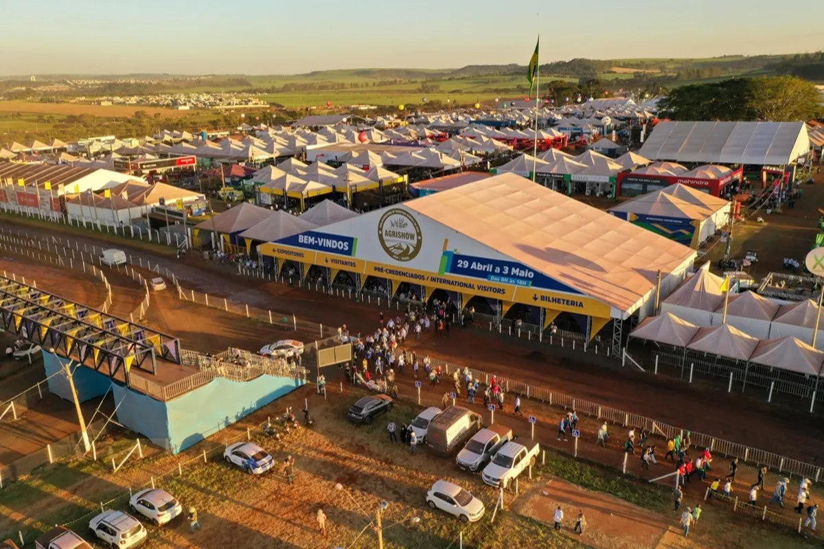 Vista aérea da entrada da feira Agrishow, com filas de visitantes, estacionamento e diversos estandes de empresas agrícolas, realizada sob tendas em amplo espaço ao ar livre.