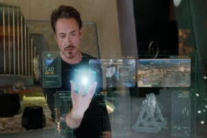 Tony Stark (Robert Downey Jr.) interagindo com hologramas 3D de dados e gráficos futuristas, segurando um objeto holográfico brilhante em sua mão.