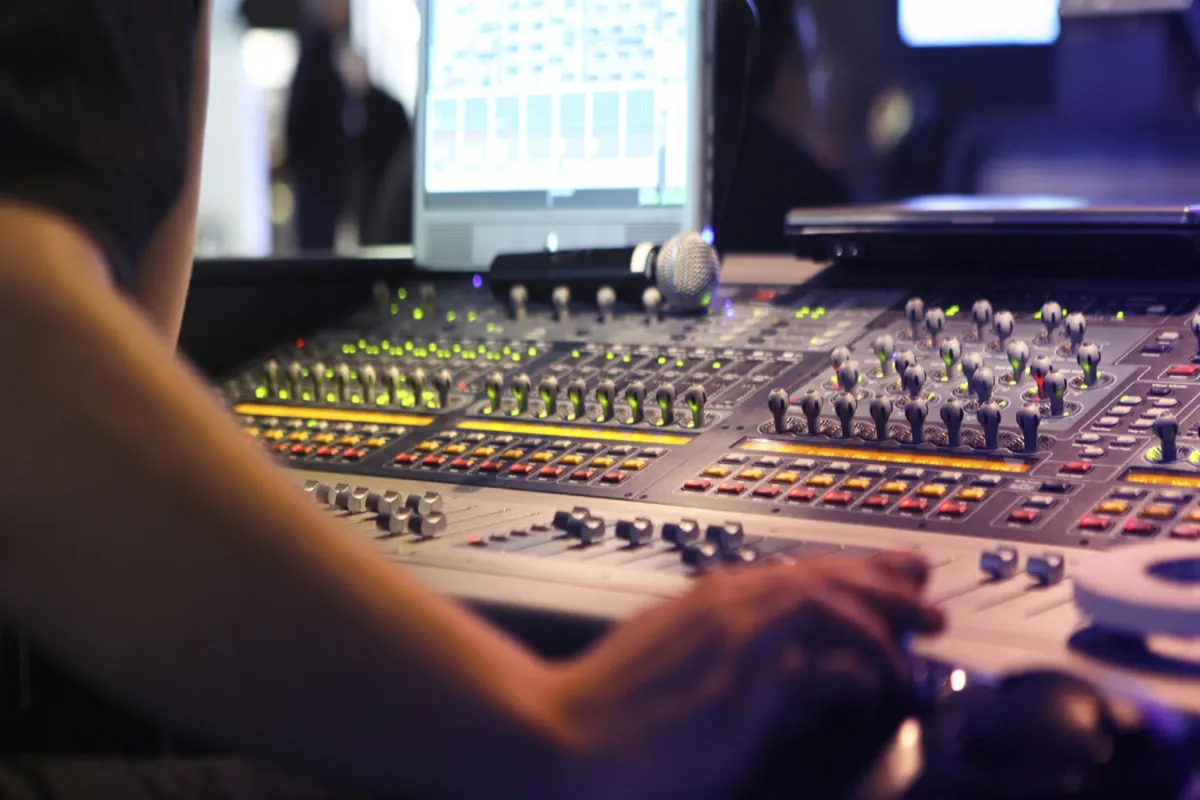 Técnico de som ajustando mesa de som estéreo profissional com luzes e botões iluminados em estúdio de gravação.