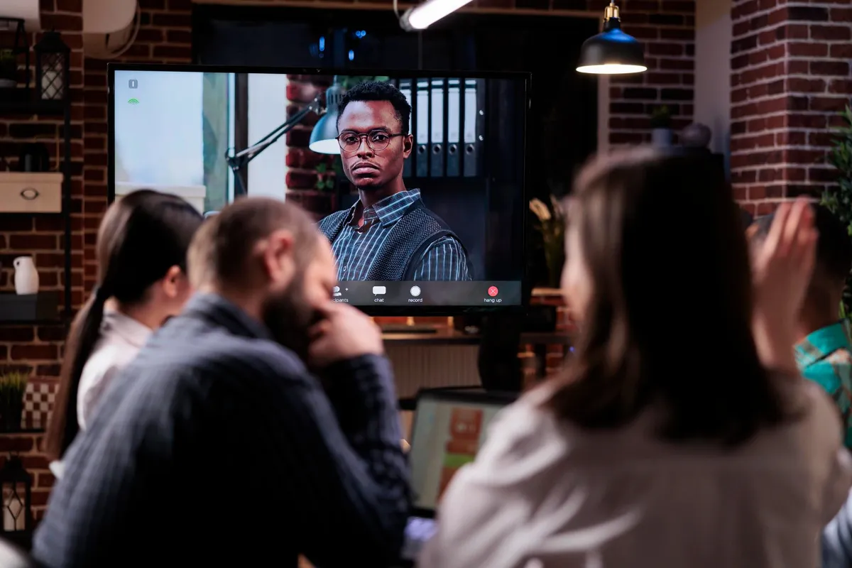 Um grupo de colegas em uma sala de reunião com paredes de tijolos assiste a uma chamada de vídeo conferência em uma tela de TV, com um homem negro em destaque na chamada.