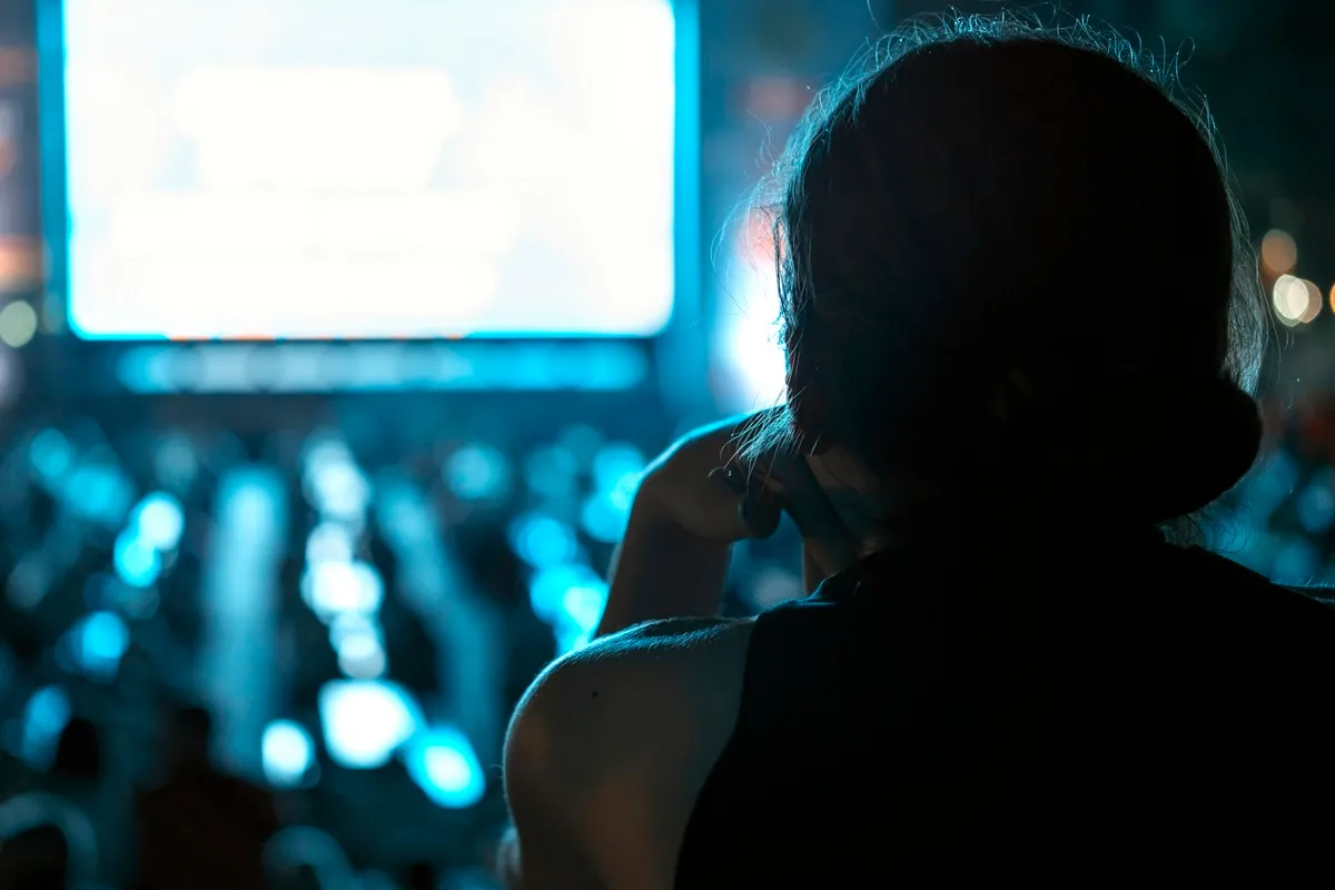 Silhueta de uma pessoa assistindo a um telão iluminado em um evento noturno com público ao fundo.