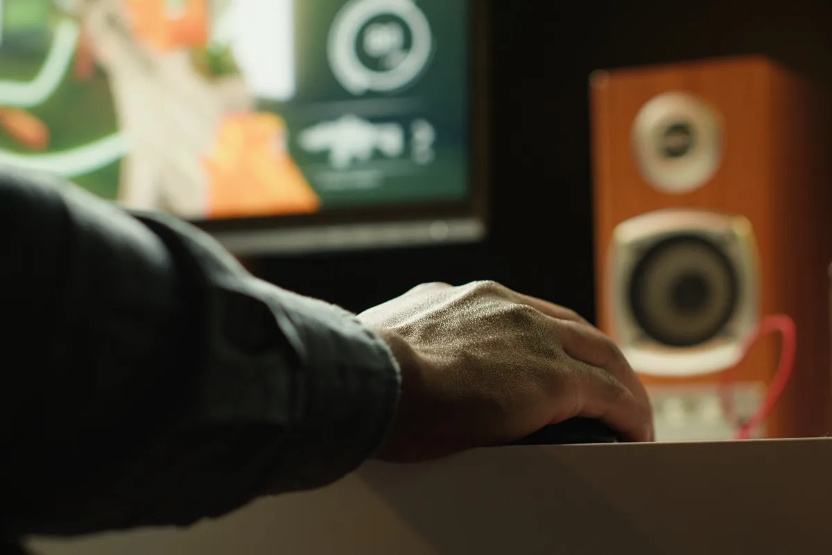 Mão de uma pessoa usando o mouse enquanto um monitor de áudio de madeira aparece desfocado ao fundo, próximo a uma tela com jogo aberto.