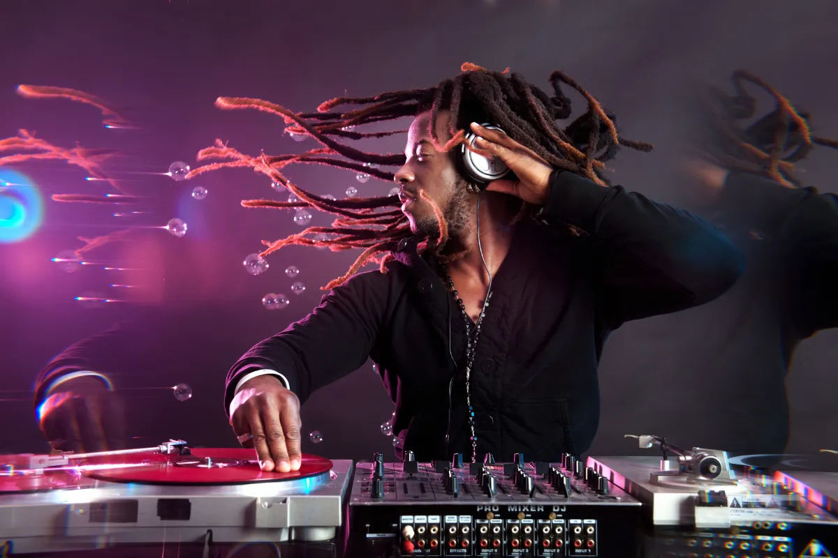 DJ com dreadlocks usando fones de ouvido enquanto toca música em uma mesa de mixagem e toca-discos em ambiente iluminado.